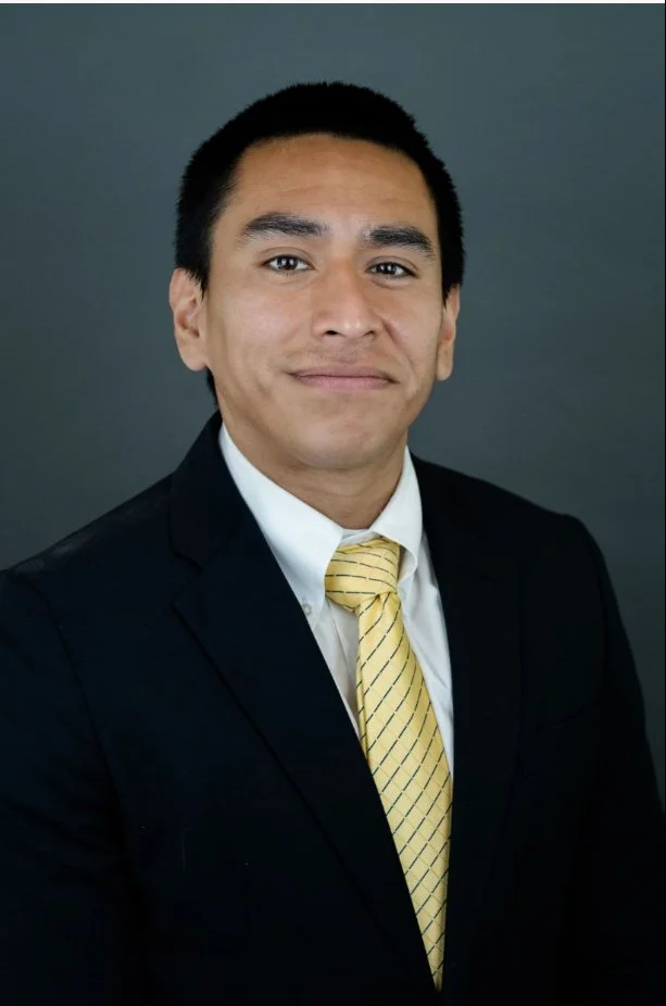 A man in a black suit, white shirt, and yellow striped tie posing against a gray background.