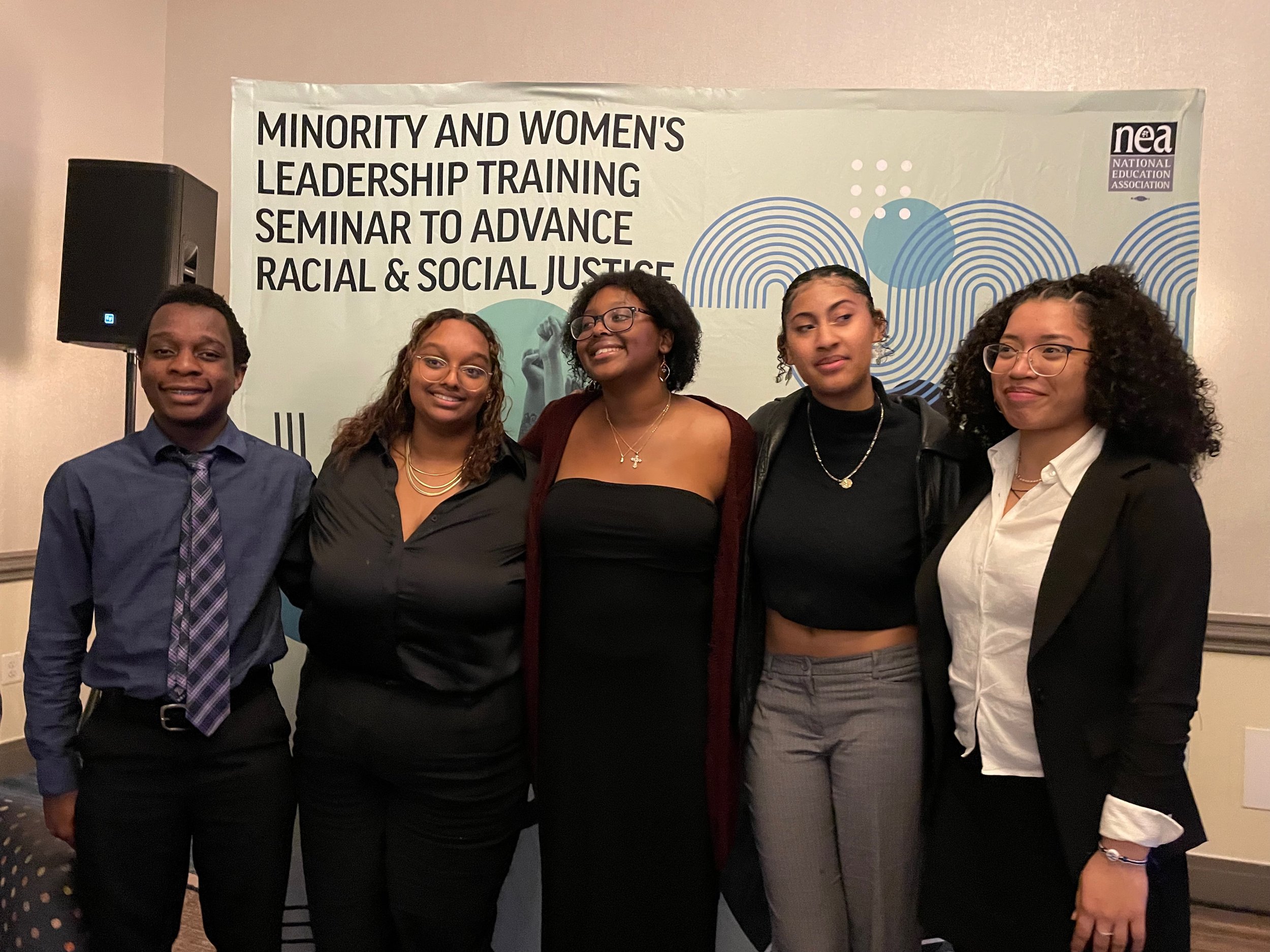 Five young women standing together in front of a banner at a seminar on racial and social justice, smiling and posing for the photo.