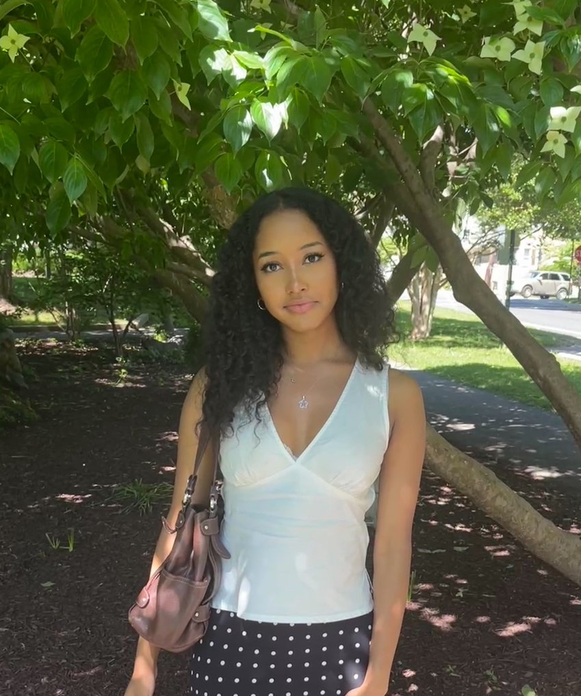 A young woman with curly black hair standing outdoors under a large leafy tree, wearing a white sleeveless top and a black polka dot skirt, holding a brown handbag, with a sidewalk and parked cars in the background.