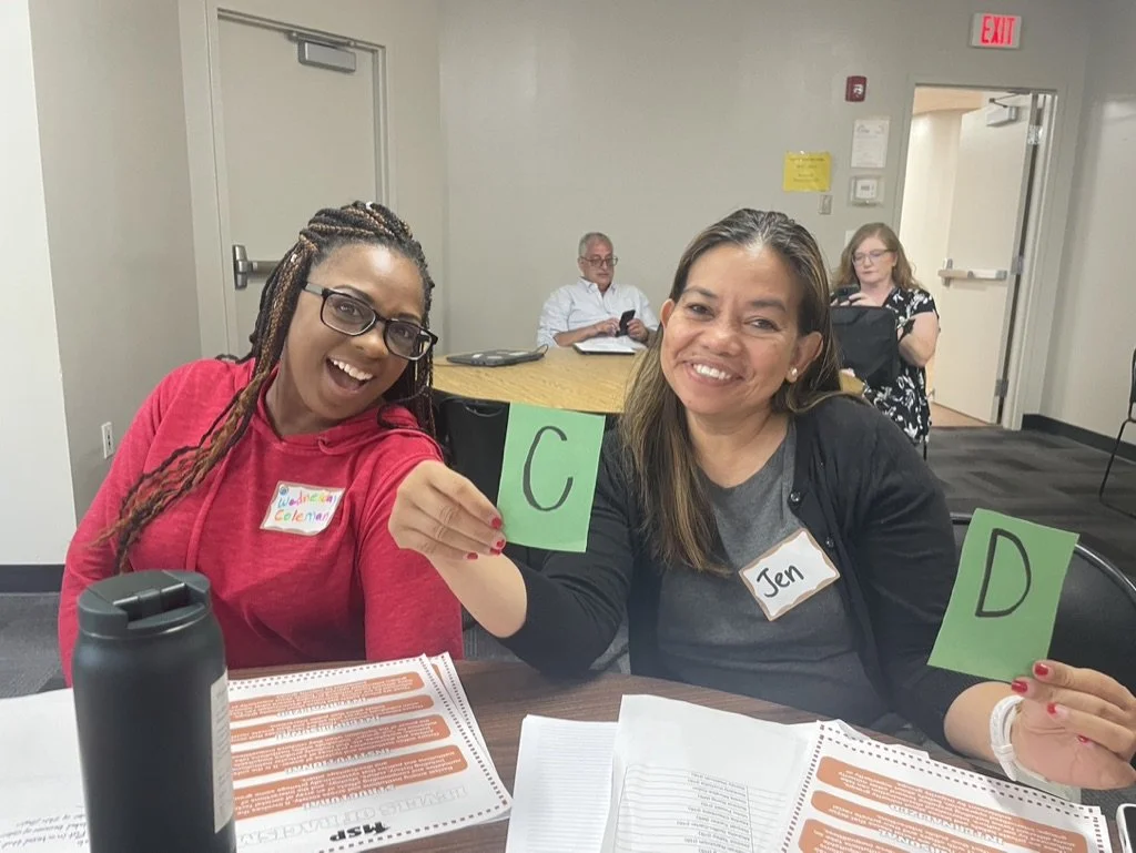 Two women sitting at a table, holding up green cards with the letters C and D, smiling. One woman wears a pink hoodie and glasses, and the other wears a dark jacket with a gold name tag reading 'Jen'. There are papers and a water bottle on the table,