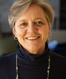 Close-up of a smiling woman with short gray hair, wearing a black turtleneck and a beaded necklace.
