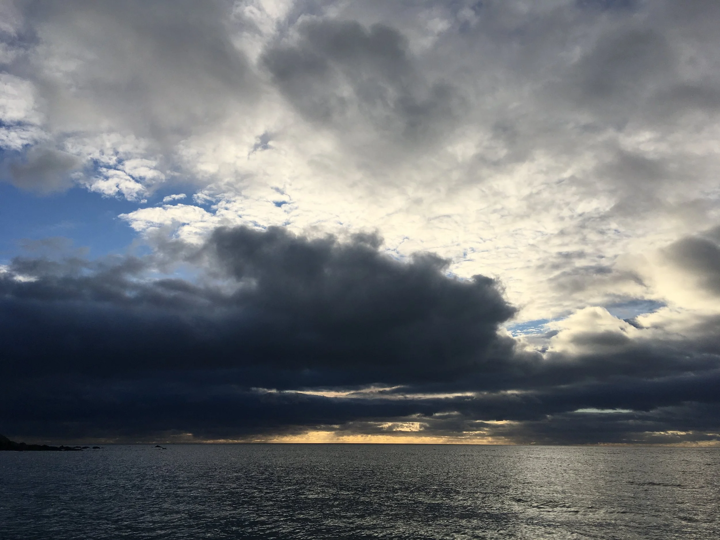 Overcast sky with dark and light clouds, calm ocean water below, with limited sunlight near the horizon.
