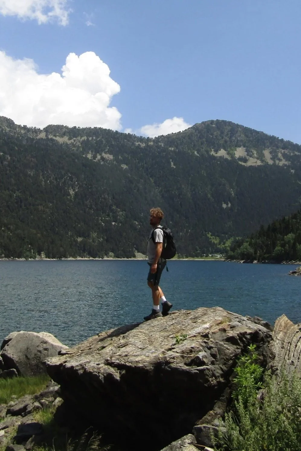 A person standing on a large rock by a lake with mountains in the background, under a partly cloudy sky.