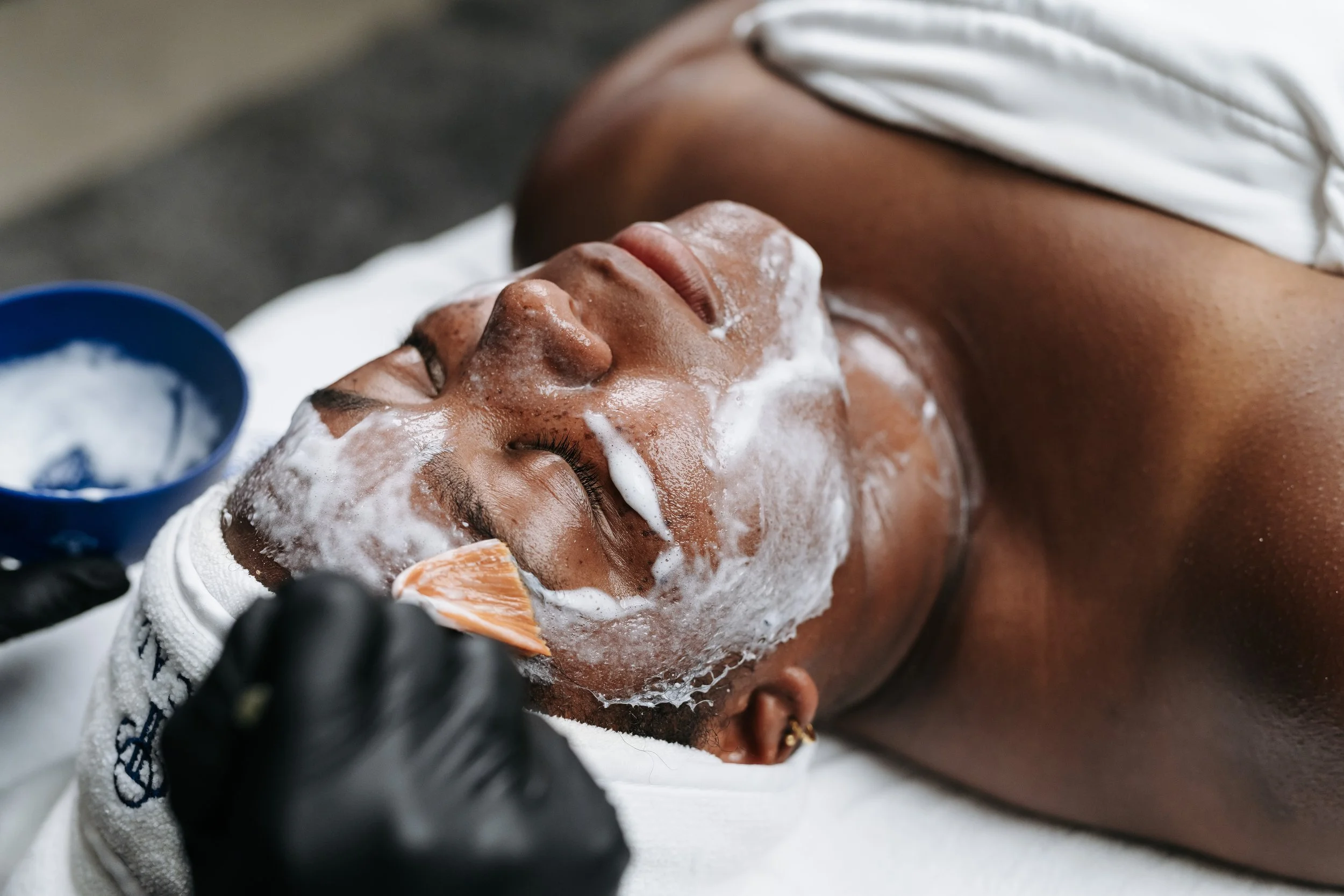 Person receiving a facial treatment, lying down with eyes closed, covered in foam, with a shower cap on head.