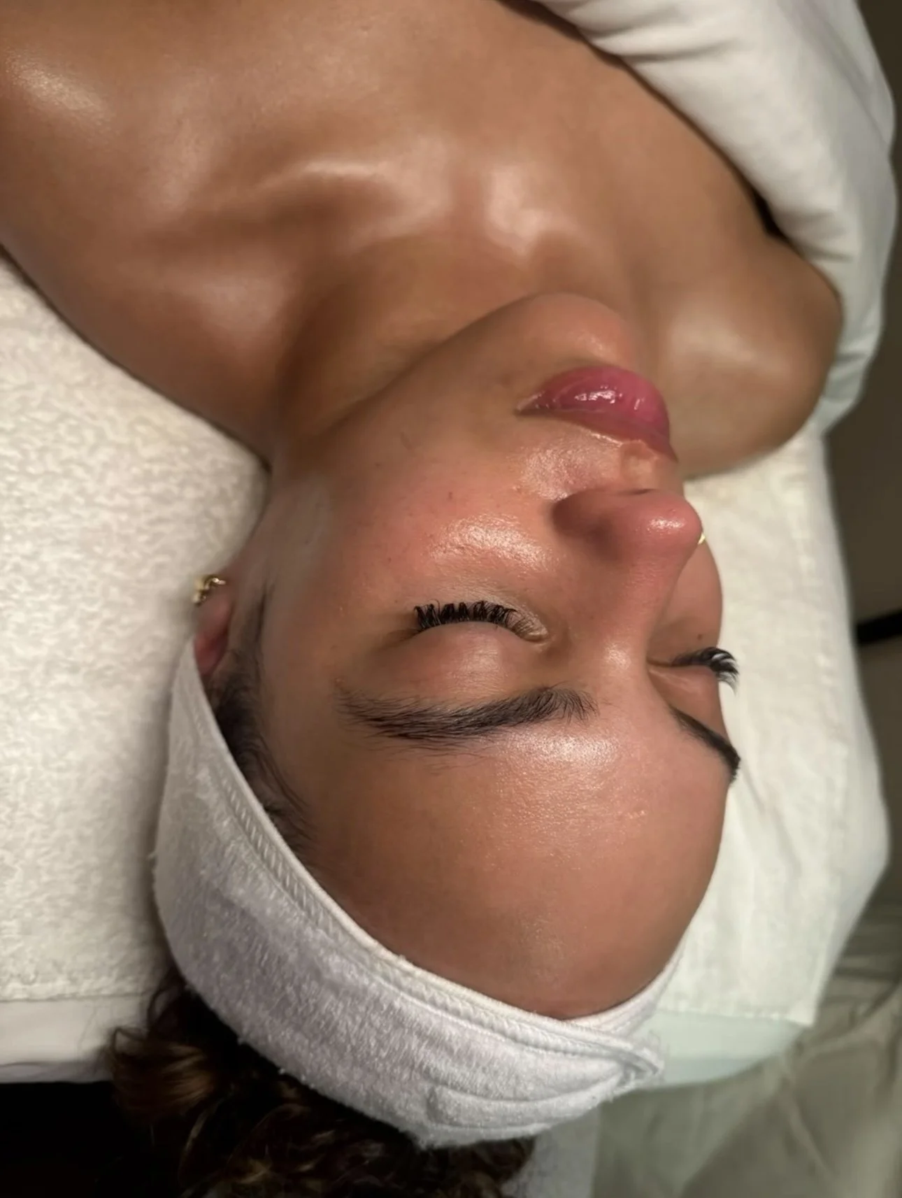 A woman with dark hair, gold earrings, and makeup lying on her back with eyes closed, wearing a white headband and towel around her head, in a spa or skincare setting.