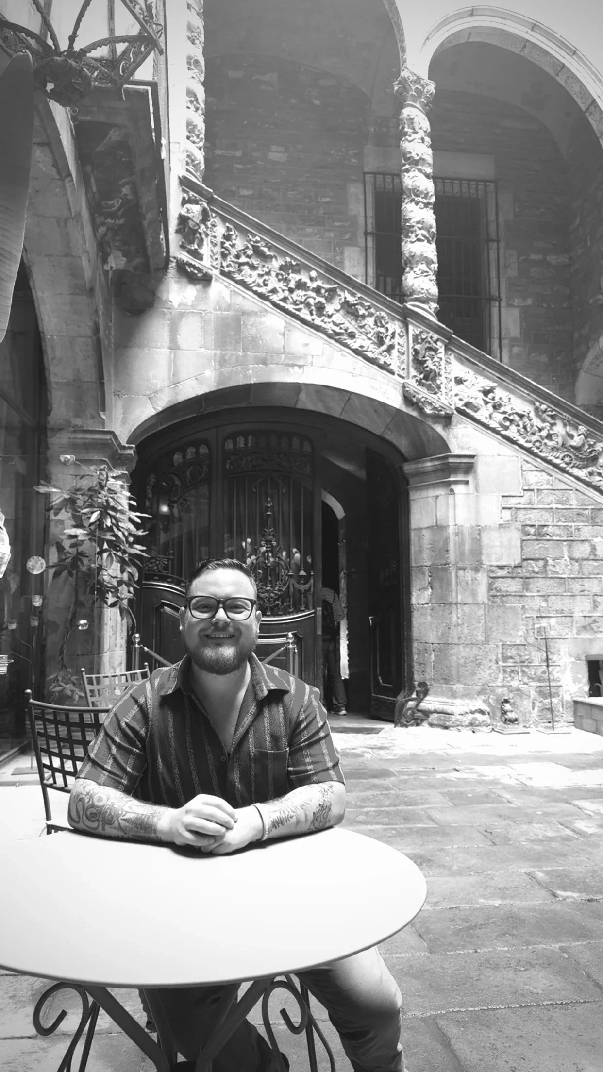 A man with glasses and a beard sitting at an outdoor table, smiling, with an ornate stone building in the background.