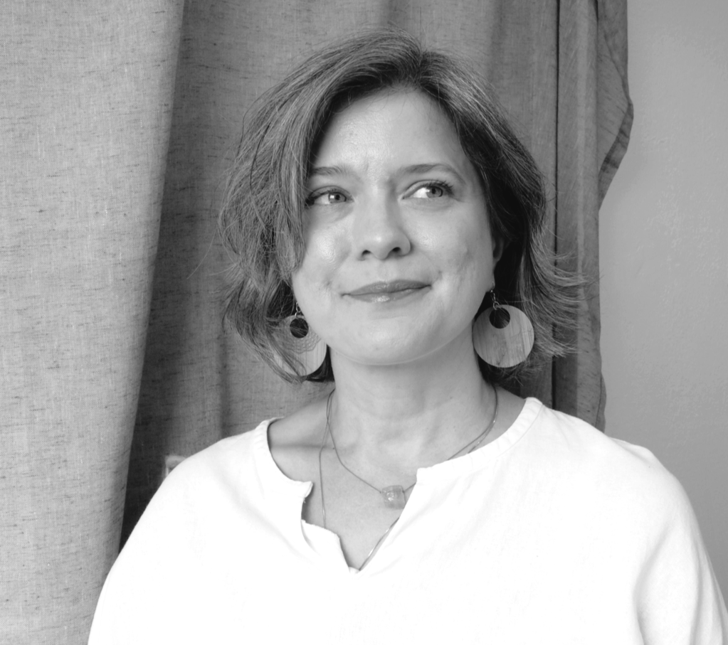 A woman with short, wavy hair wearing large earrings and a light-colored blouse, standing in front of a curtain and a wall, looking to her right with a slight smile.