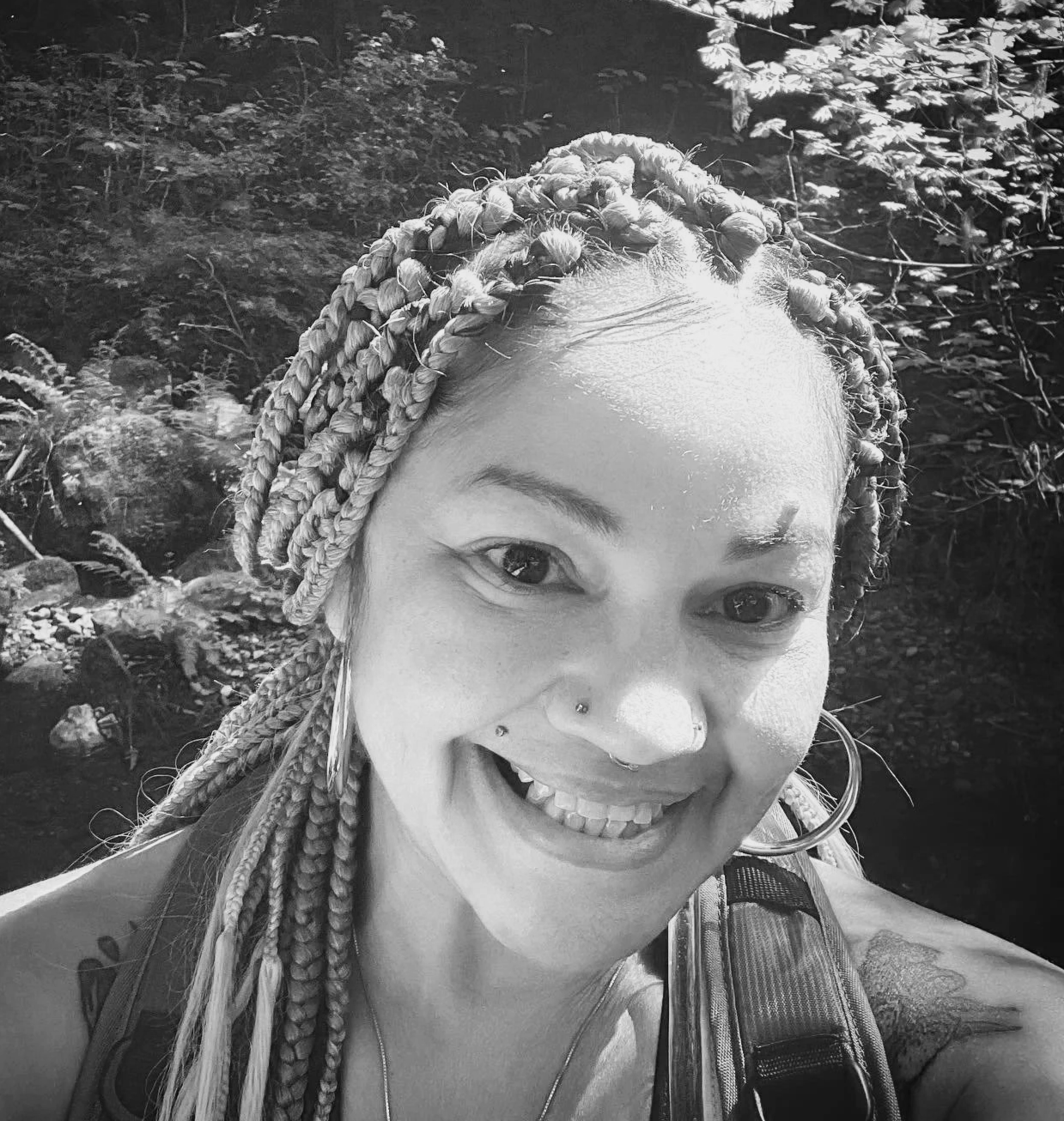 A smiling woman with braided hair and facial piercings, outdoors near a stream or river, in black and white.