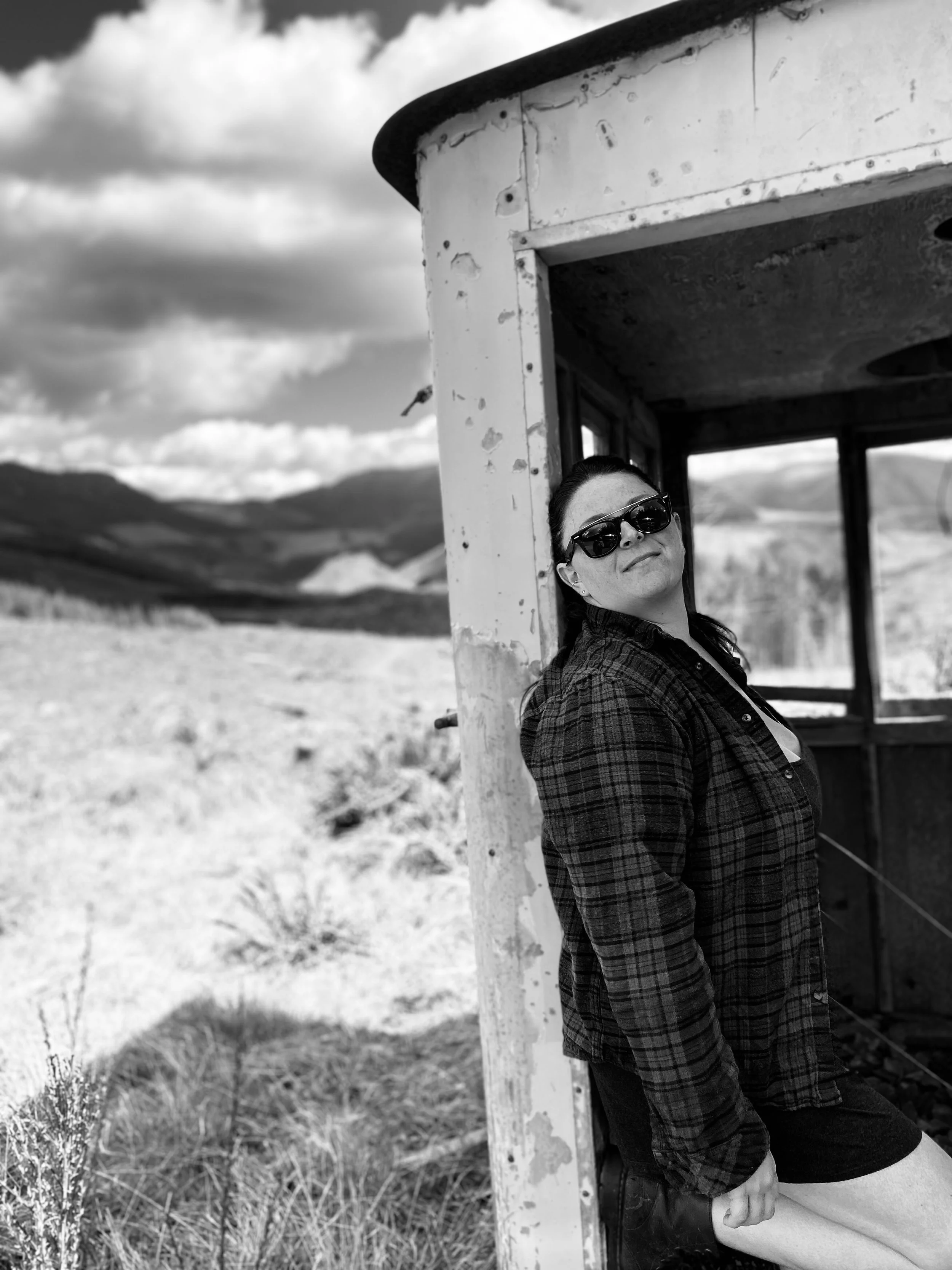 A woman wearing sunglasses and a plaid shirt leaning against the corner of an old, weathered train caboose in a rural landscape with hills and clouds in the background.