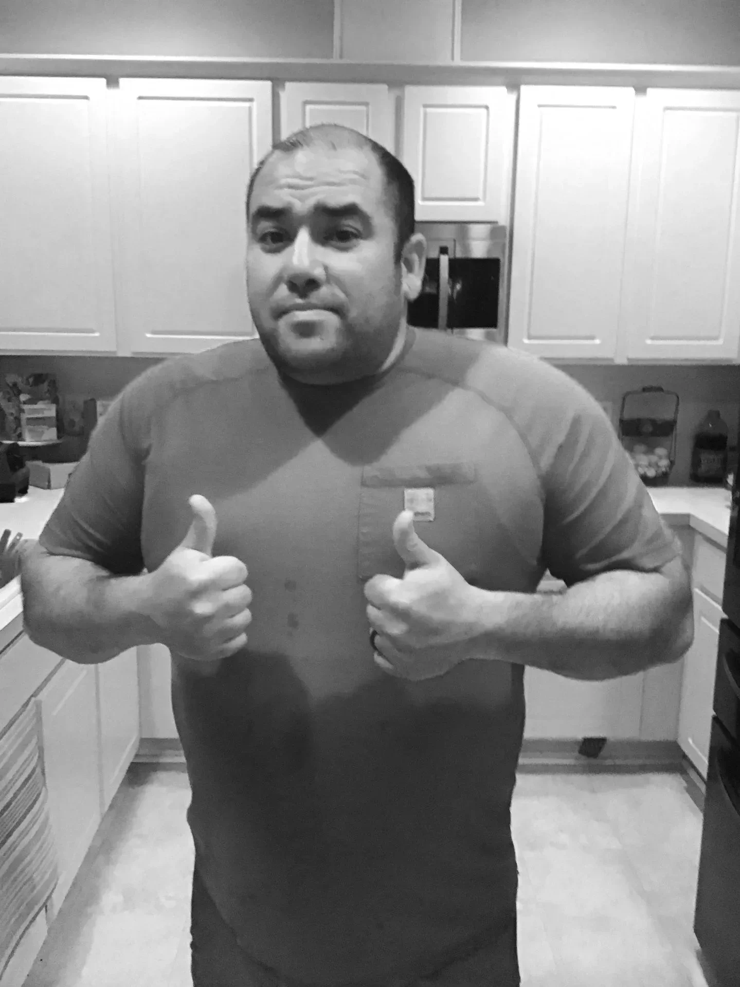 A man gives a thumbs-up gesture with both hands in a kitchen, wearing a casual t-shirt, in a black and white photo.