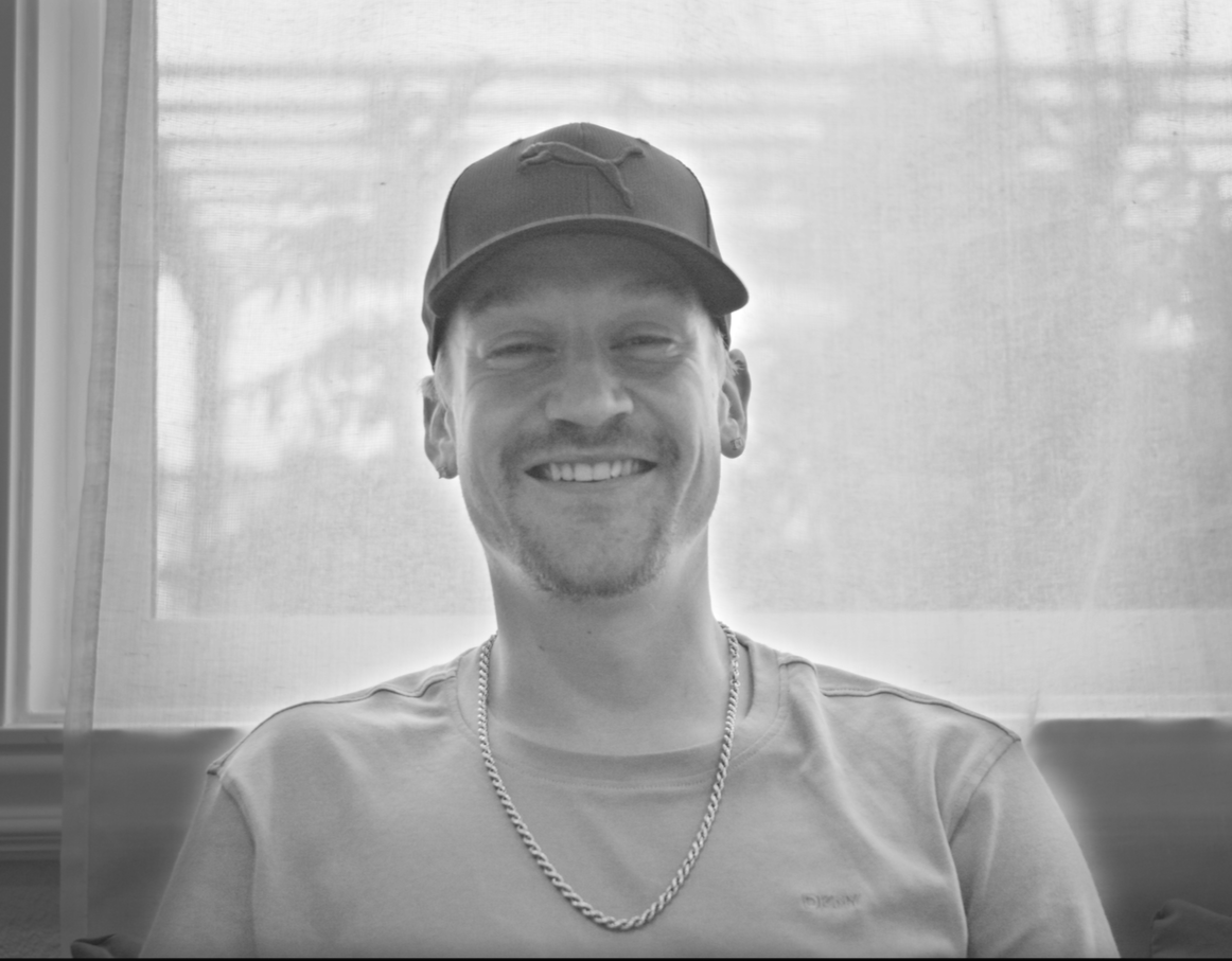 A young man with a big smile, wearing a baseball cap, chain necklace, and a T-shirt, sitting in front of a window with curtains.