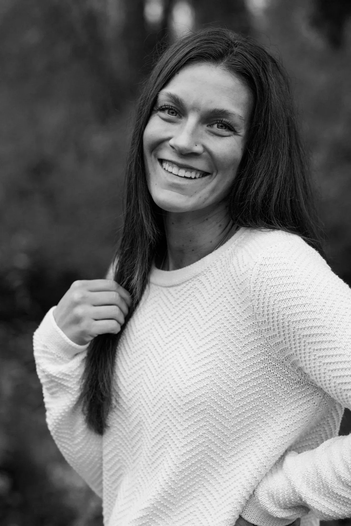 Black and white portrait of a woman smiling outdoors, with long hair, wearing a textured sweater, with a blurred natural background.