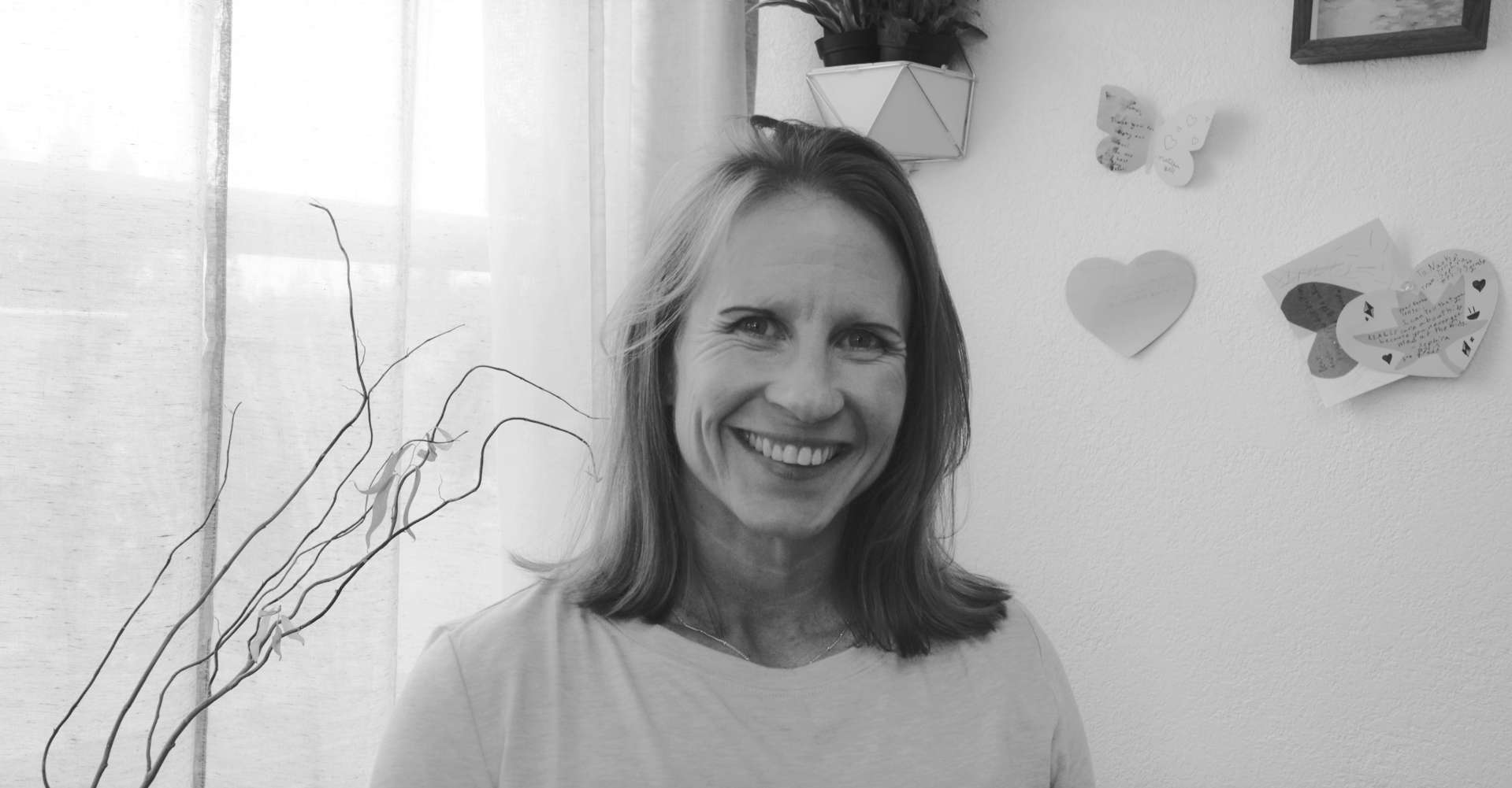 A woman smiling in front of a wall decorated with paper hearts and notes, with a window and some plants in the background.