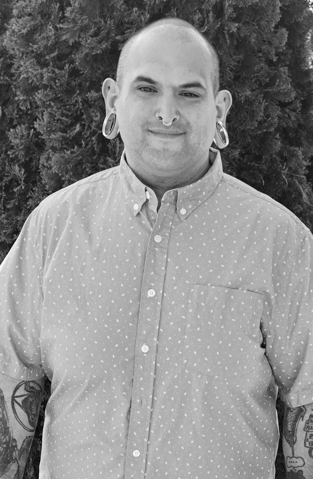 Black-and-white photo of a person with large gauges, short hair, and tattoos on both arms, wearing a patterned button-up shirt, standing outdoors with bushes in the background.