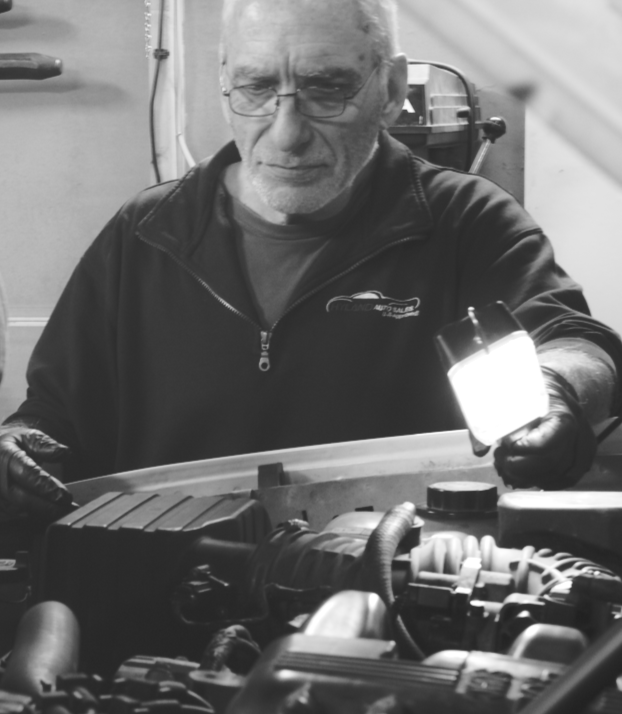 An older man with glasses inspecting a car engine in a garage, holding a flashlight.