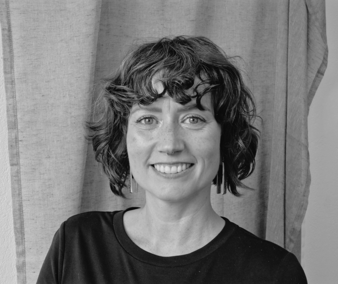 Black and white photo of a woman with short, curly hair, smiling, wearing earrings and a dark shirt, standing in front of a curtain.