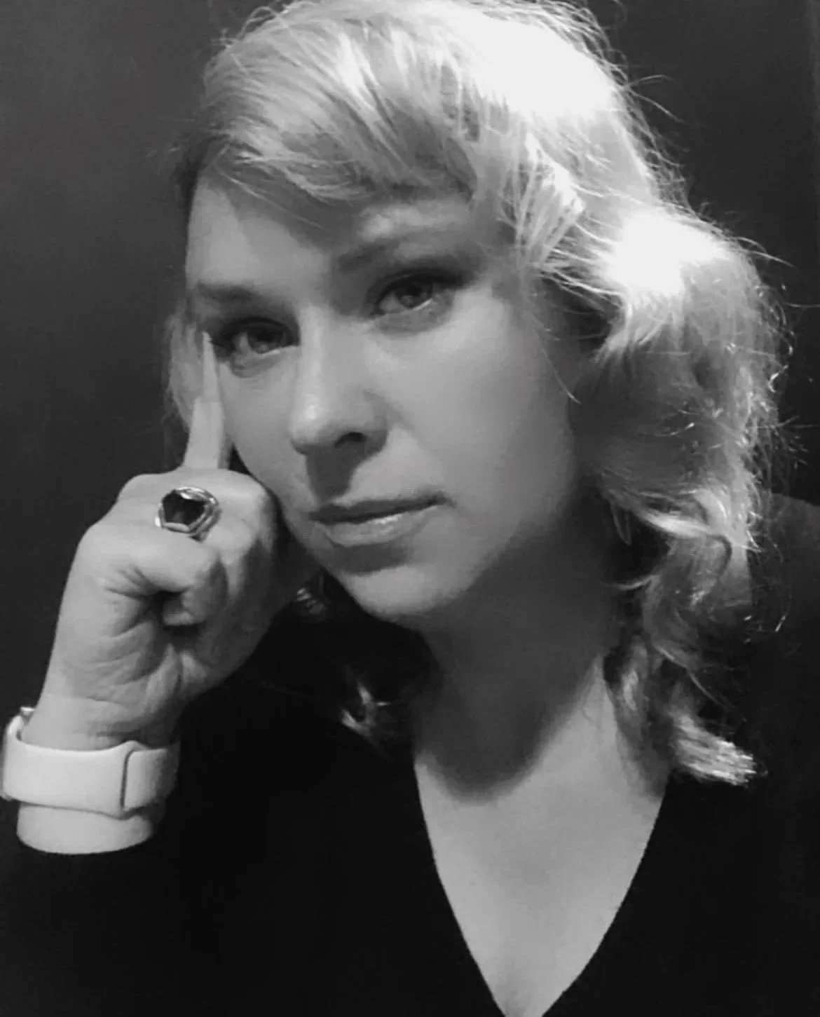 Black and white portrait of a woman with wavy blonde hair, wearing a watch, a ring, and a dark top, posing with her finger touching her temple.