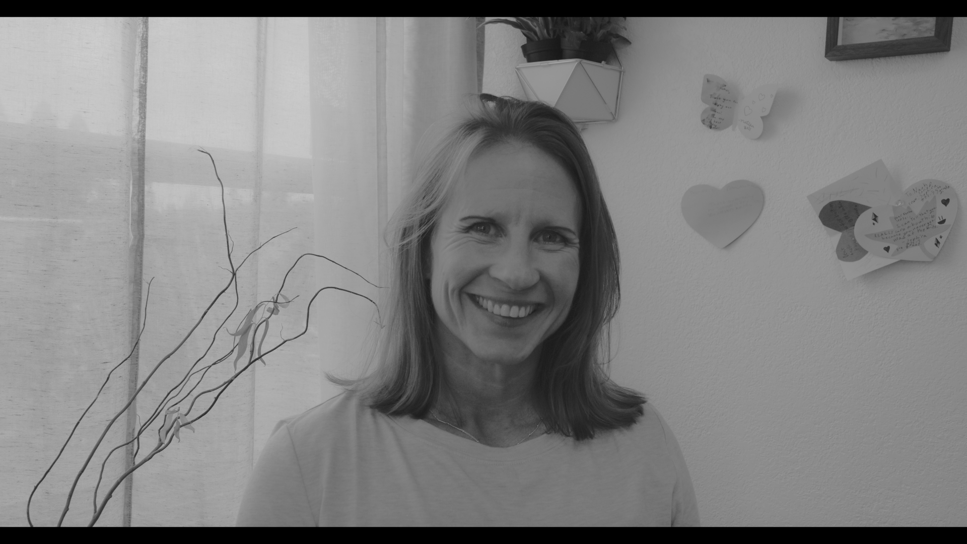 A woman smiling in front of a wall decorated with paper hearts and notes, with a window and some plants in the background.