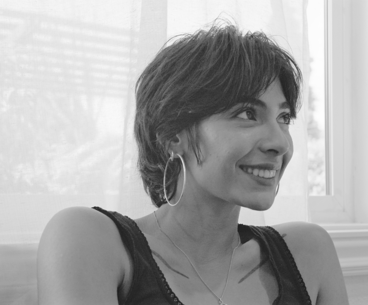 A woman with short dark hair smiling, wearing earrings and a necklace, sitting near a window with sheer curtains.