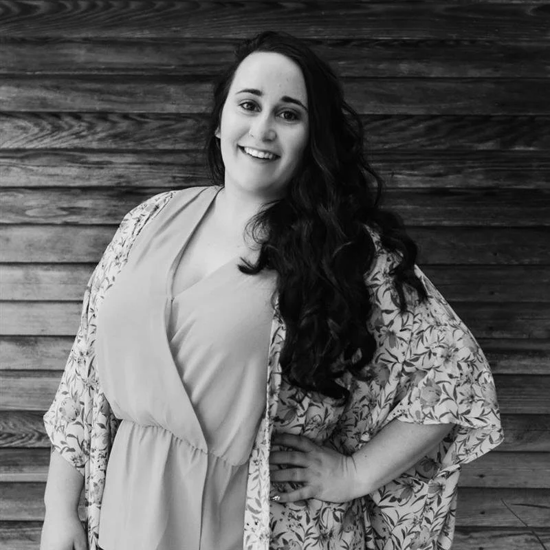 A woman with long dark curly hair, smiling, standing with one hand on her hip in front of a wooden wall, wearing a floral kimono over a light-colored dress.