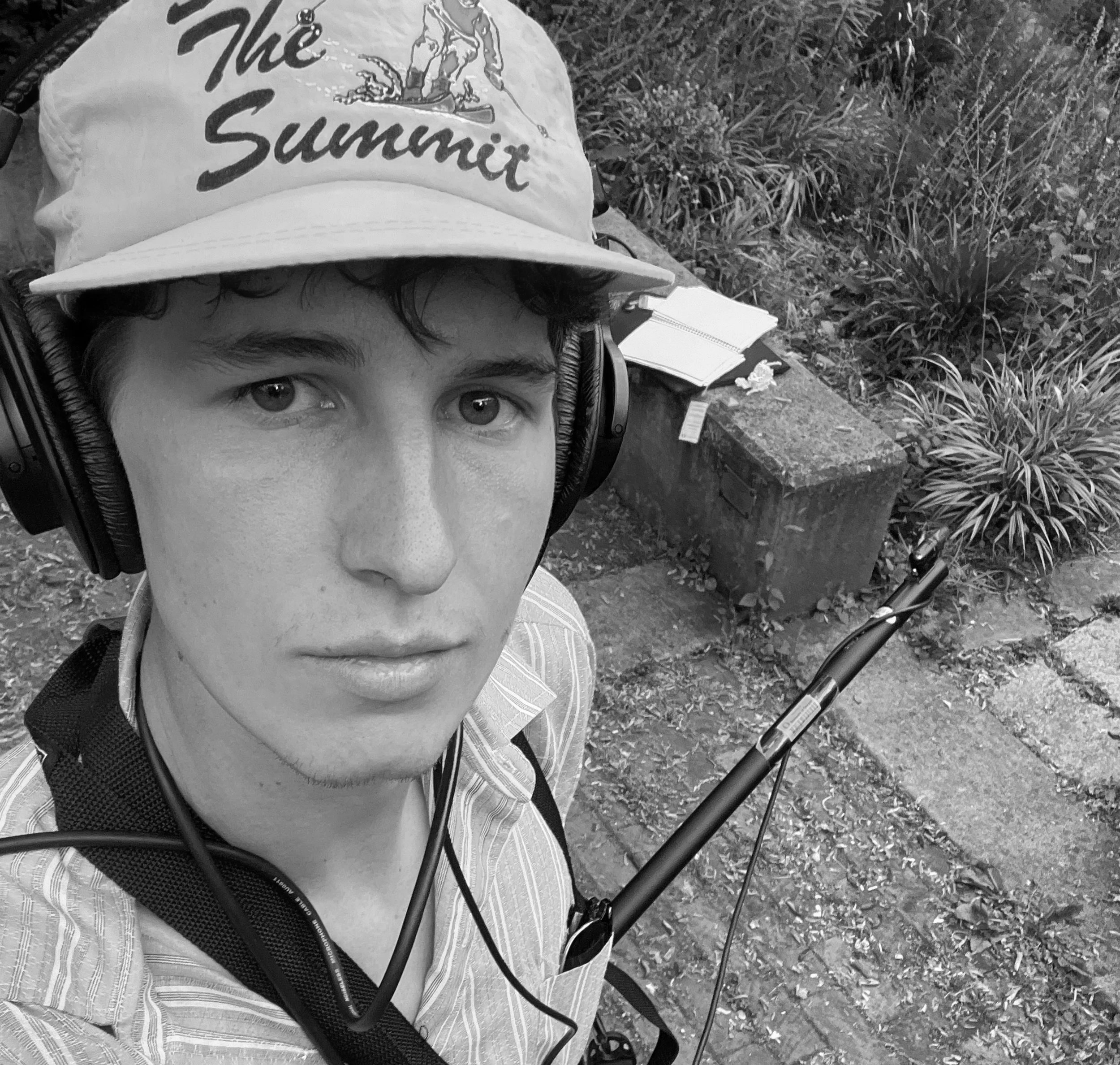A young man wearing a hat with 'The Summit' written on it, headphones, and a striped shirt taking a selfie outdoors next to a park bench and plants.