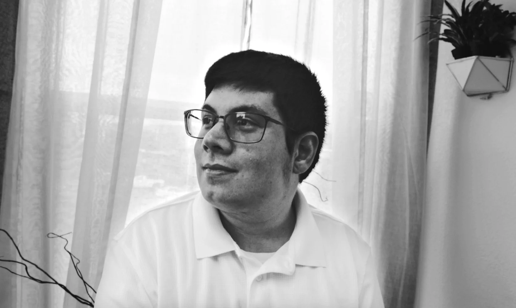 A young man with glasses and short dark hair is sitting near sheer curtains, looking to his left, with a potted plant on the wall behind him.