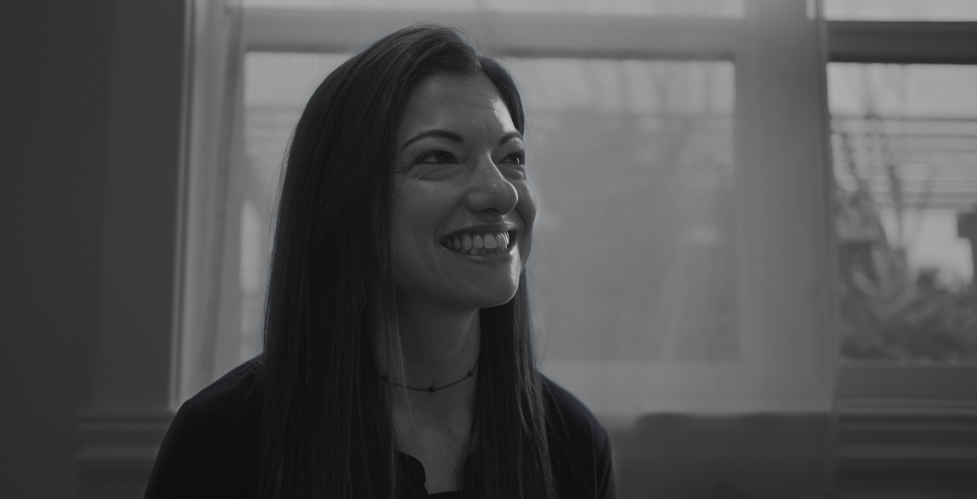 A smiling woman with long dark hair sitting in front of a window with blinds.