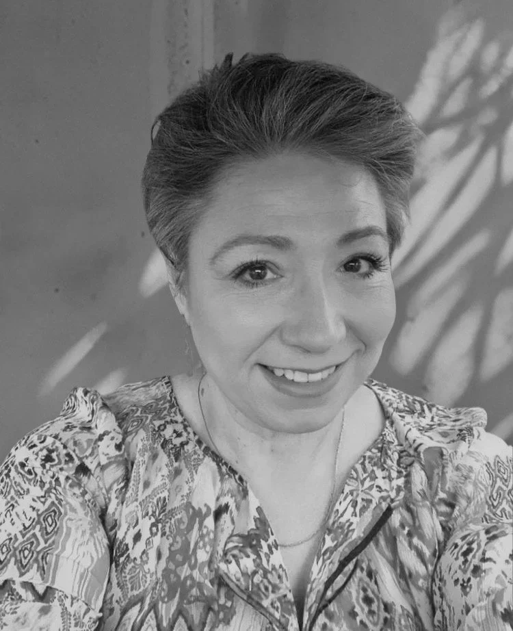 Black and white photo of a woman with short hair smiling, wearing a patterned top, standing indoors with a plant shadow on the wall behind her.