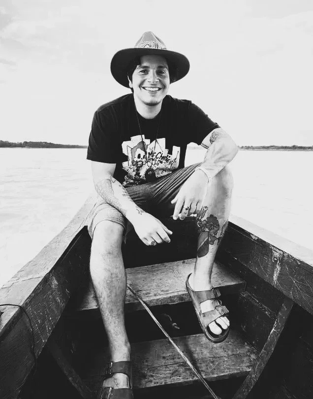 A smiling young man wearing a sombrero hat, a graphic T-shirt, shorts, and sandals sitting in a small wooden boat on a body of water, with a distant shoreline in the background.