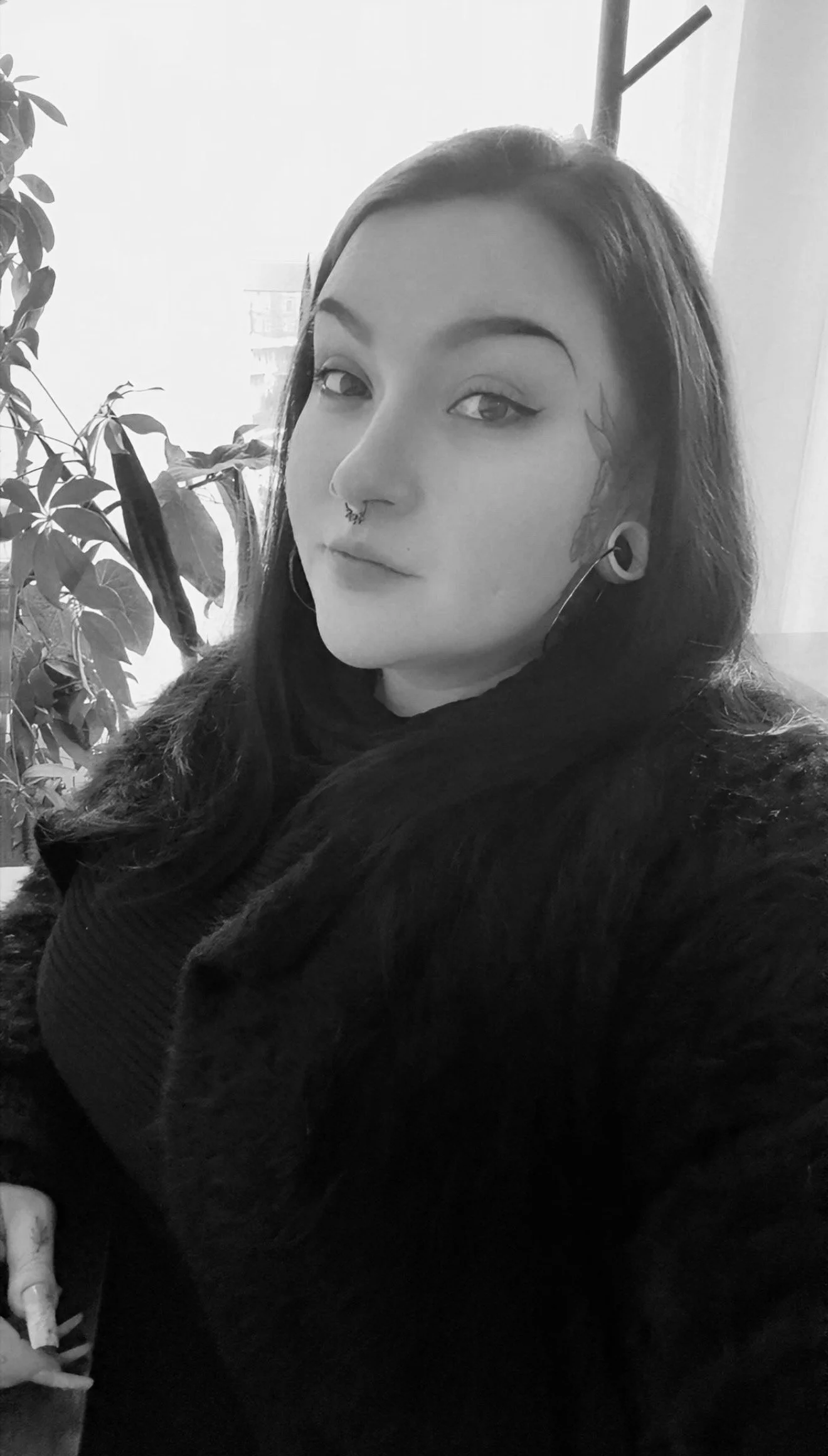 Black and white photo of a woman with dark hair, a septum piercing, ear gauges, and facial tattoos, sitting near a window and a houseplant.