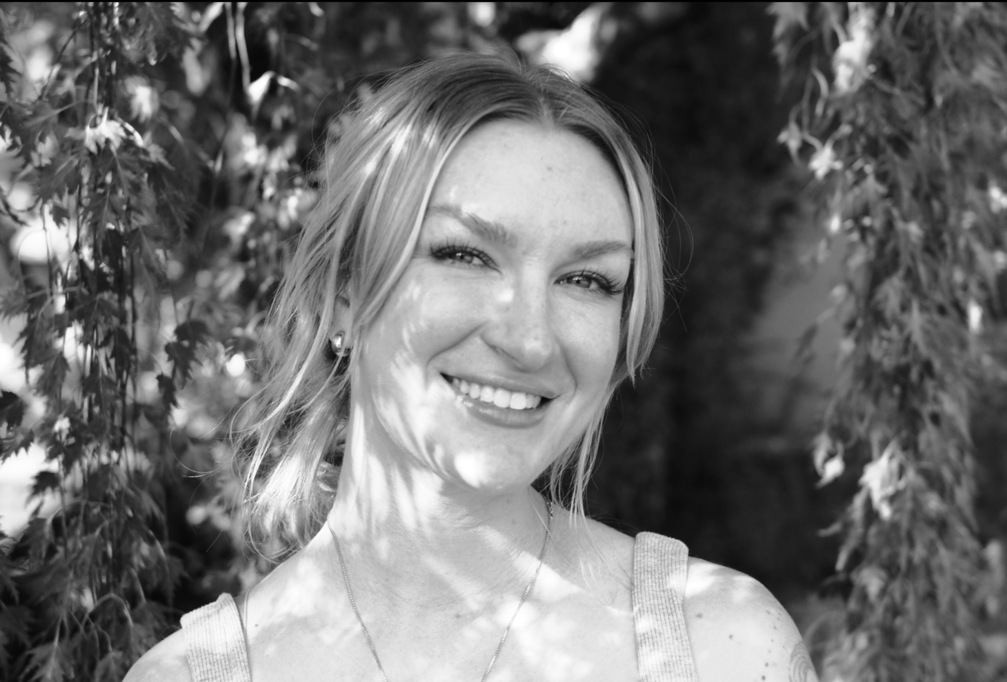 Black and white photo of a woman with shoulder-length hair smiling outdoors, with foliage and trees in the background.