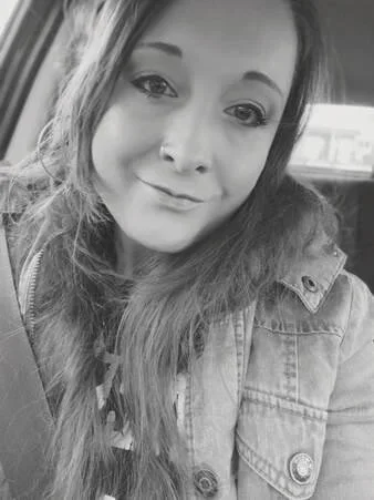 Black and white photo of a young woman with long hair, smiling slightly, wearing a denim jacket, sitting in a vehicle.