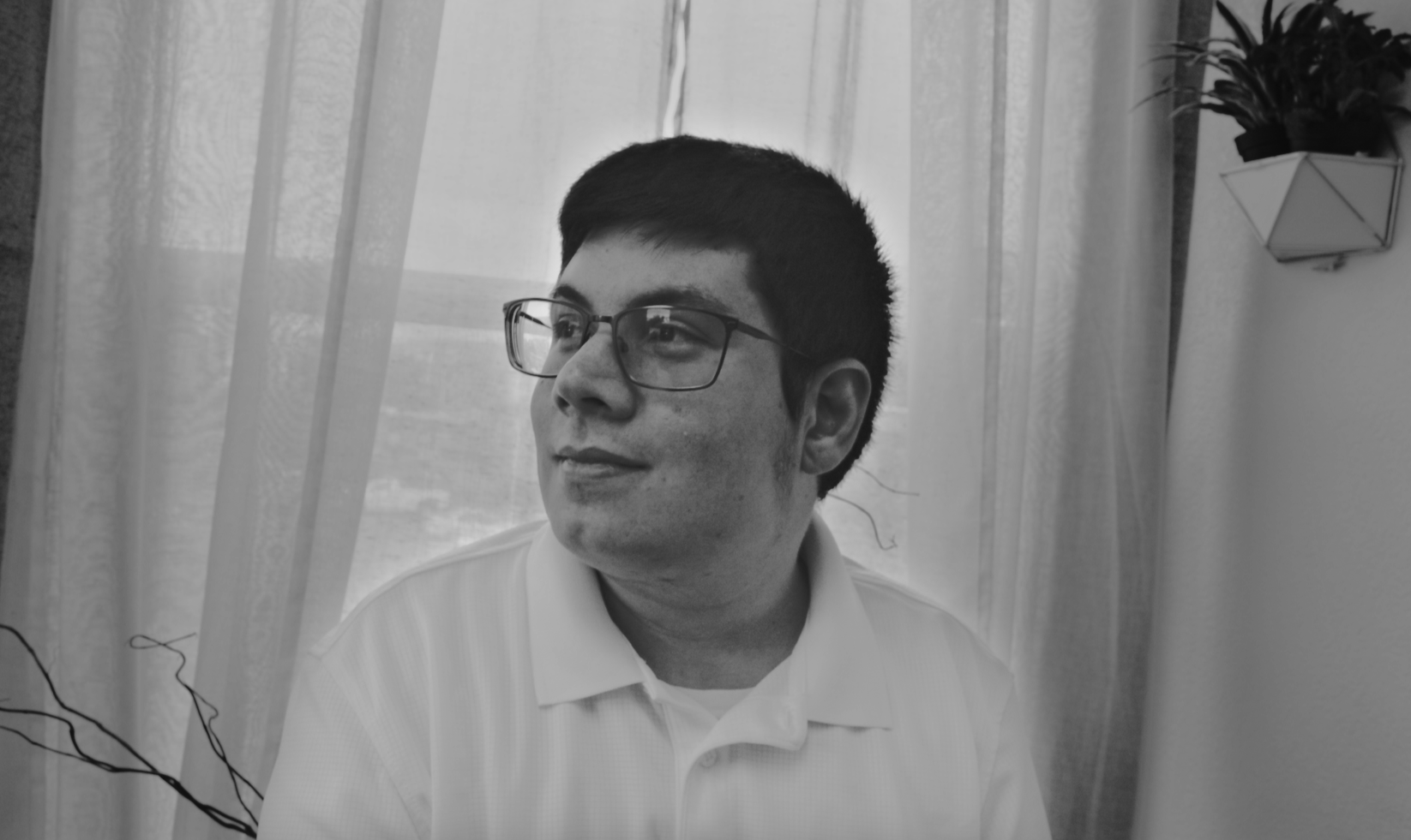A young man with glasses and short dark hair is sitting near sheer curtains, looking to his left, with a potted plant on the wall behind him.