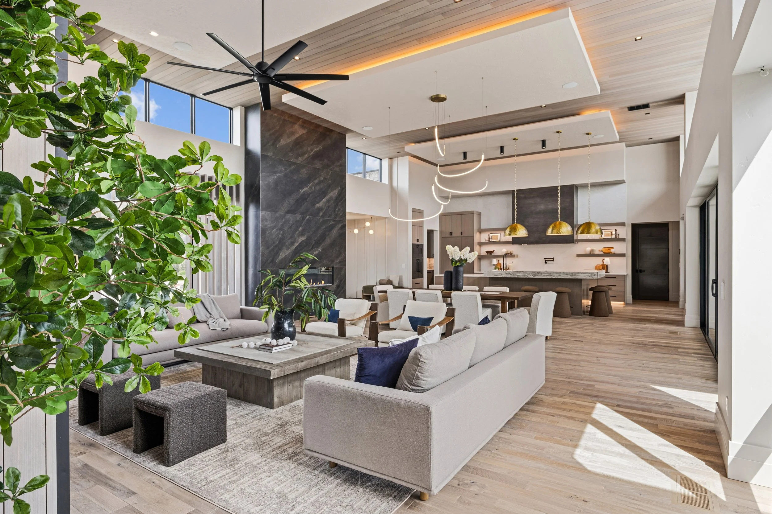 Modern open-concept living room and kitchen with large windows, wooden floors, stylish furniture, gold pendant lights, and contemporary decor.