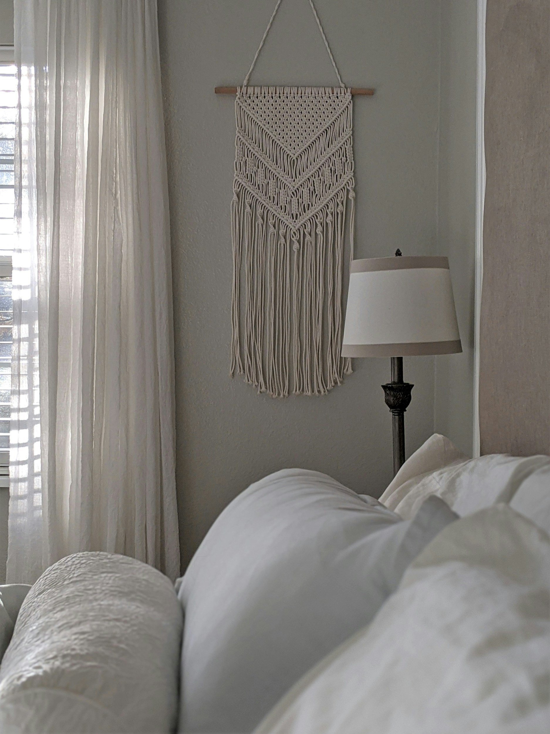 white macrame hanging on a wall by a white drape with a lampa and pillow in the foreground