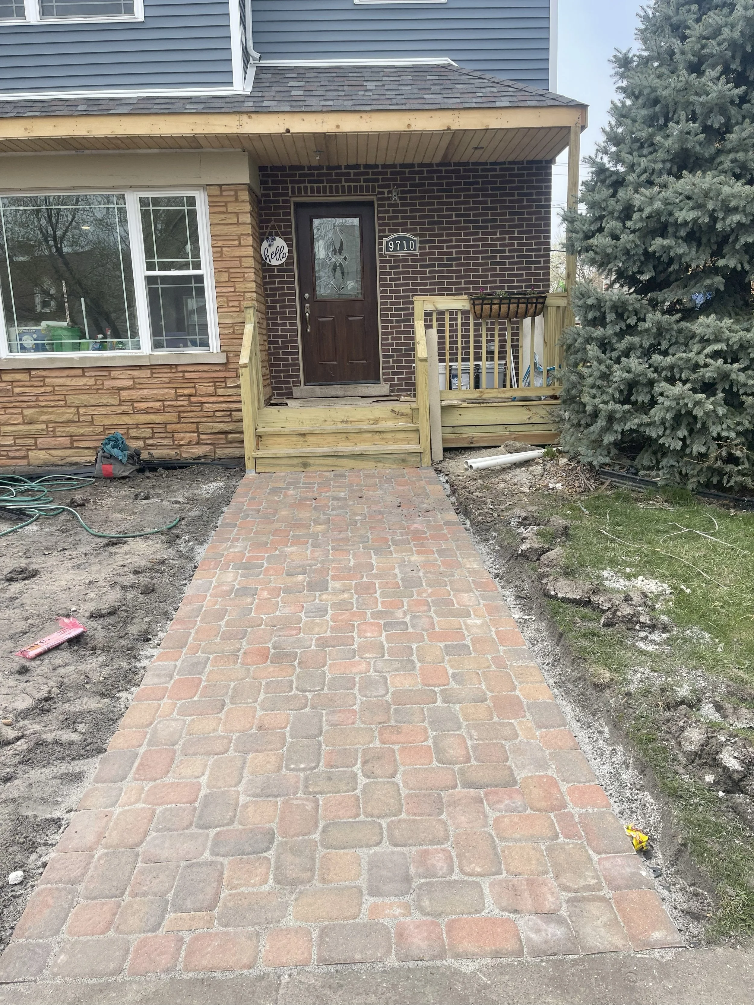 New brick walkway leading to the front door of a house under construction, with a small wooden staircase, a large window on the left, and a large evergreen tree on the right.
