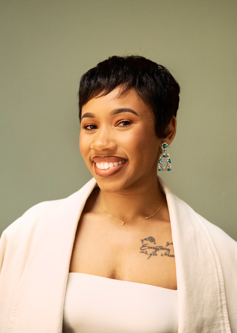 A woman with short dark hair smiling and wearing earrings, a necklace, and a cream-colored blazer over a white top, standing against a plain green background.