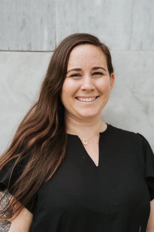 A young woman with long brown hair and a black blouse, smiling and standing outside against a light-colored wall.