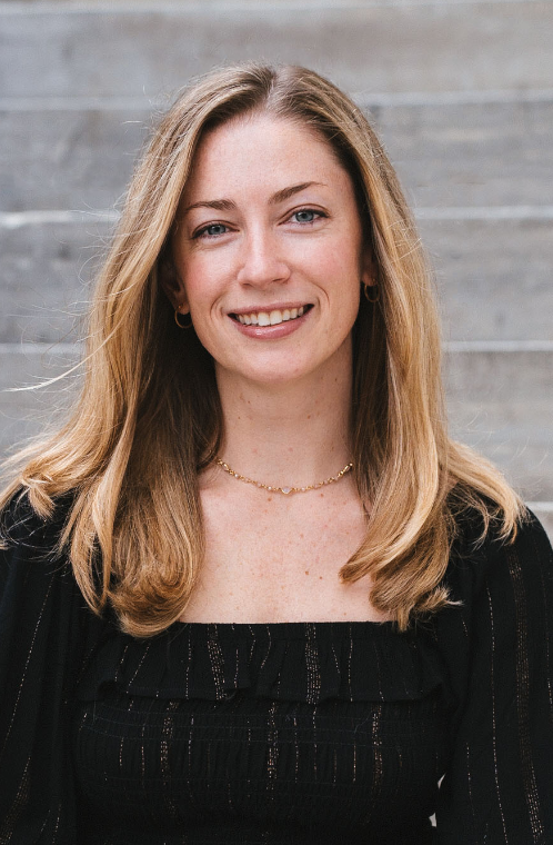 A woman with shoulder-length blonde hair, smiling, wearing a black top, gold earrings, and a gold necklace, standing in front of a wooden background.