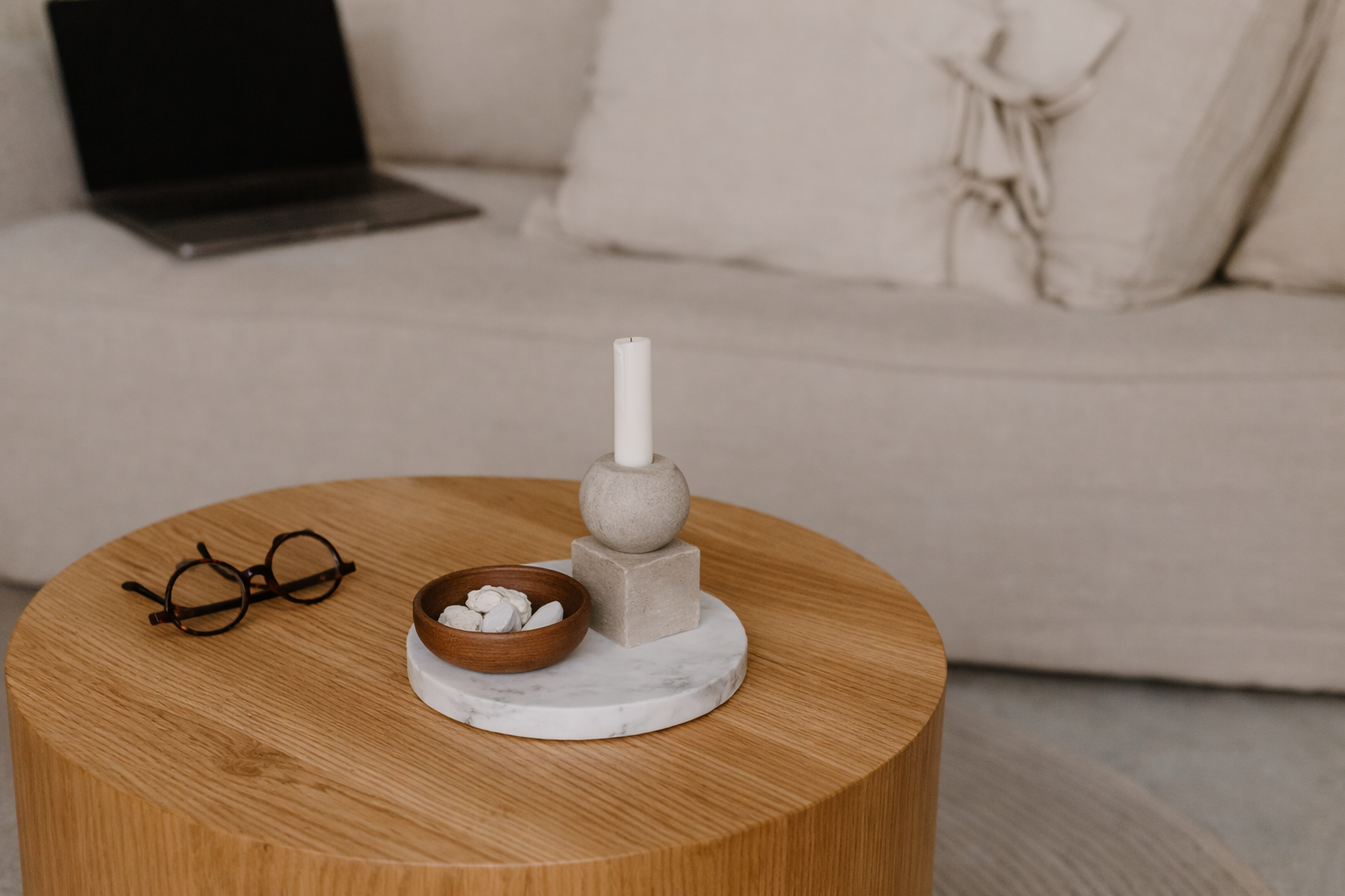Round wooden side table with a small bowl of stones, a marble base with a concrete sphere and white candle, and a pair of glasses on it, in a cozy living room.