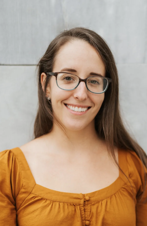 A young woman with long brown hair, wearing glasses and a mustard-colored shirt, smiling in front of a gray tiled wall.