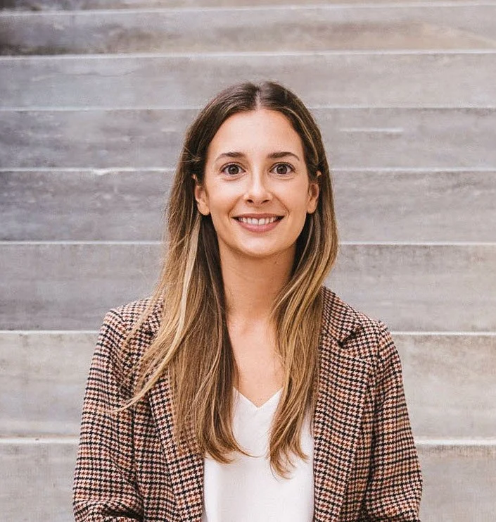 A woman with long light brown hair, smiling, wearing a plaid blazer and white top standing on gray concrete stairs.
