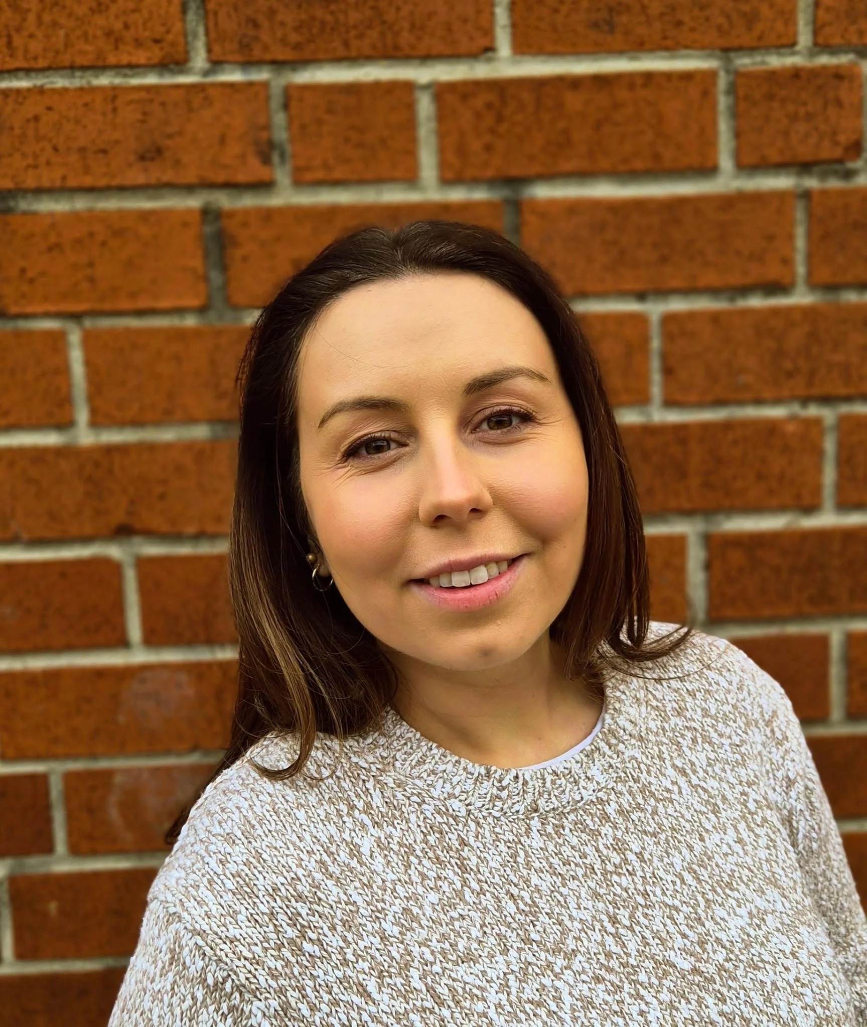 A woman with shoulder-length brown hair smiling in front of a red brick wall.