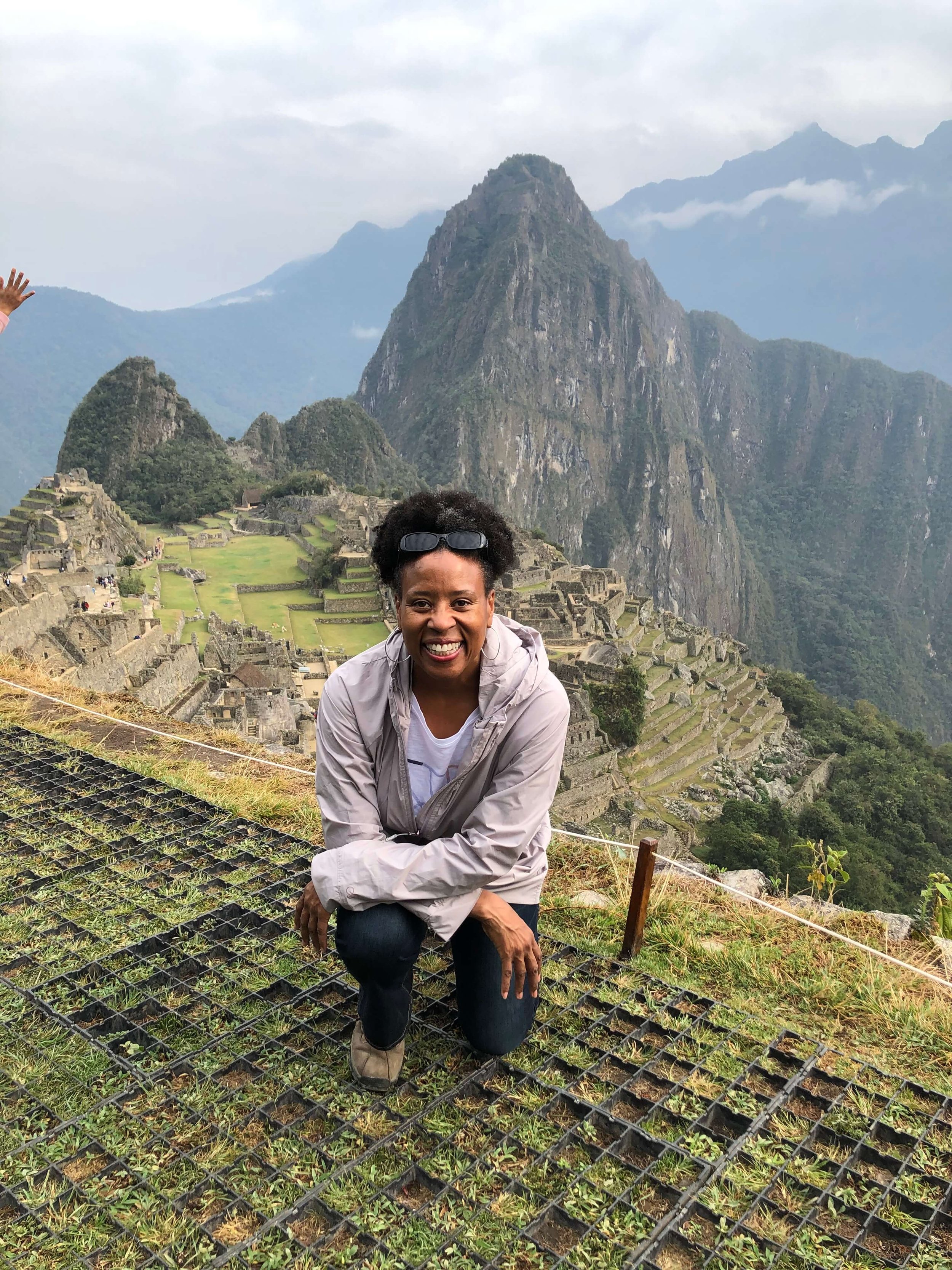 Pamela Oakes  smiling and crouching on a grassy area with Machu Picchu and mountain peaks in the background.