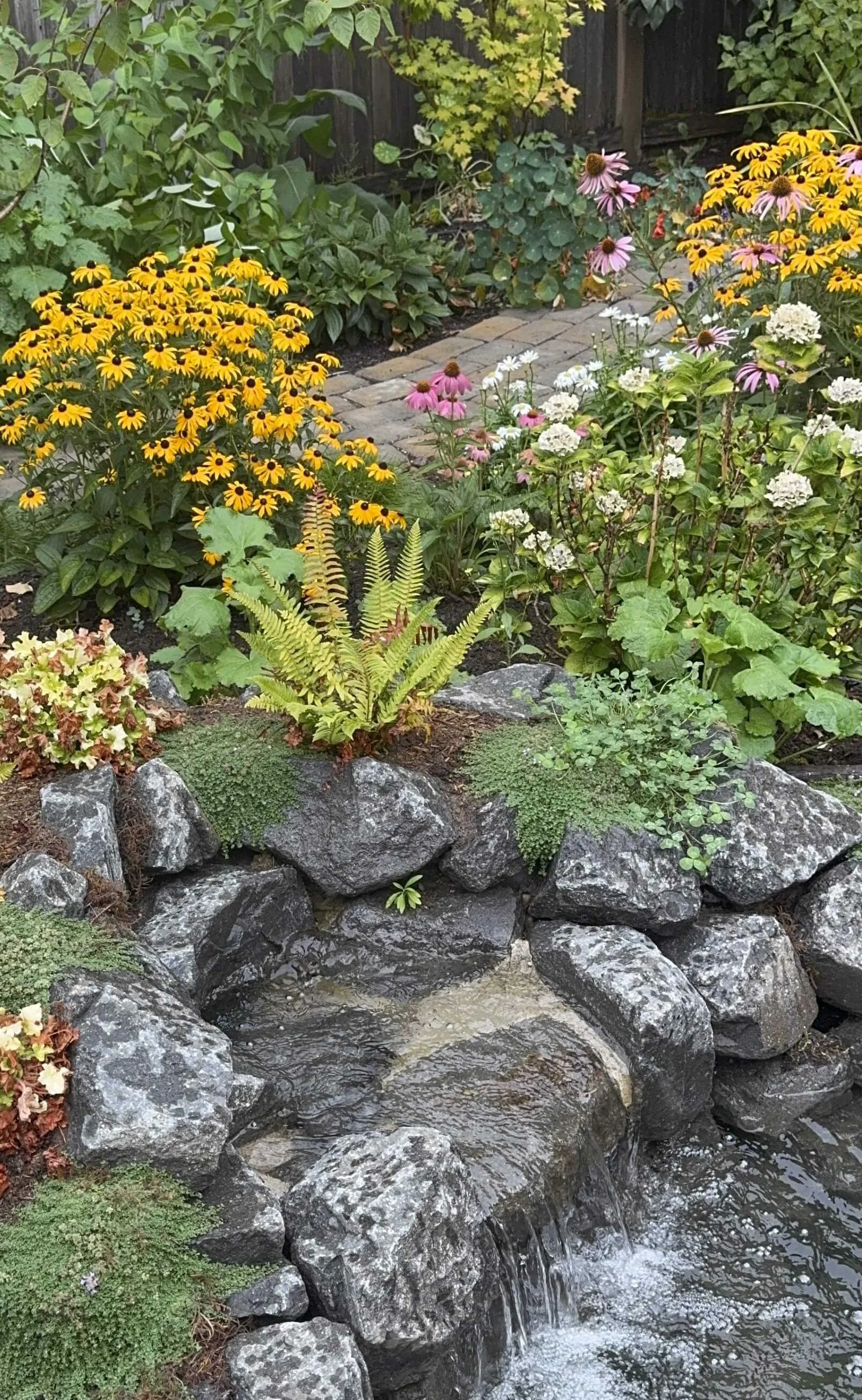 A lush garden with yellow, pink, white, and peach flowers surrounding a small, rocky waterfall with water flowing over the rocks, and a brick pathway visible in the background.