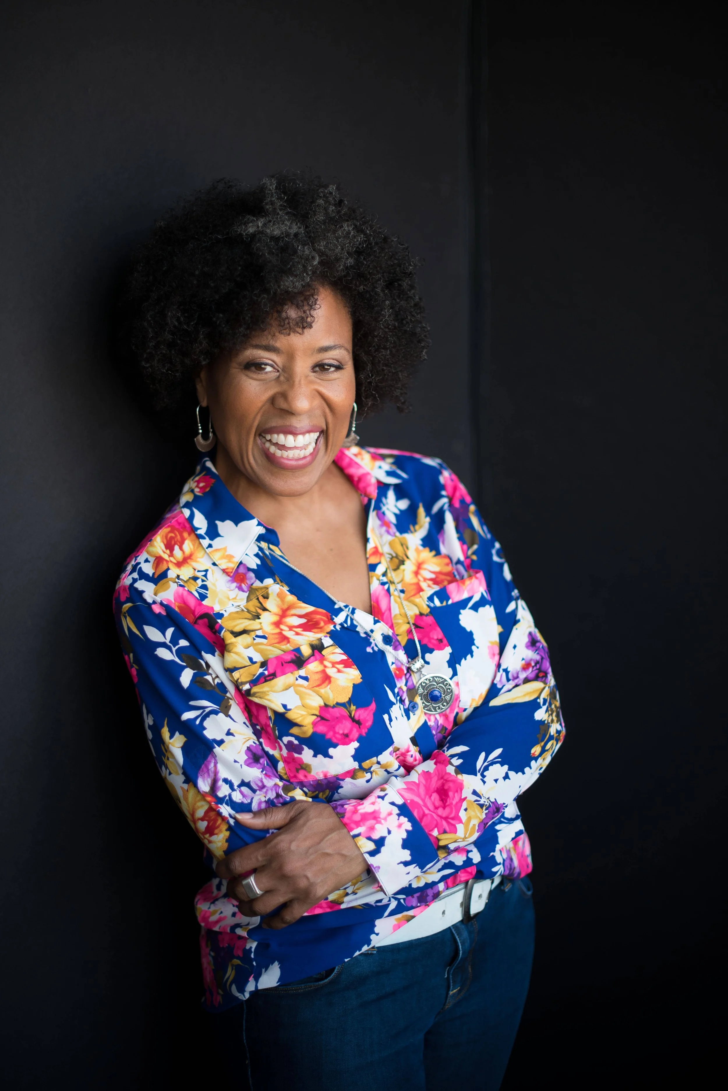 Pamela Oakes  wearing a vibrant floral blouse and jeans, standing against a black background.