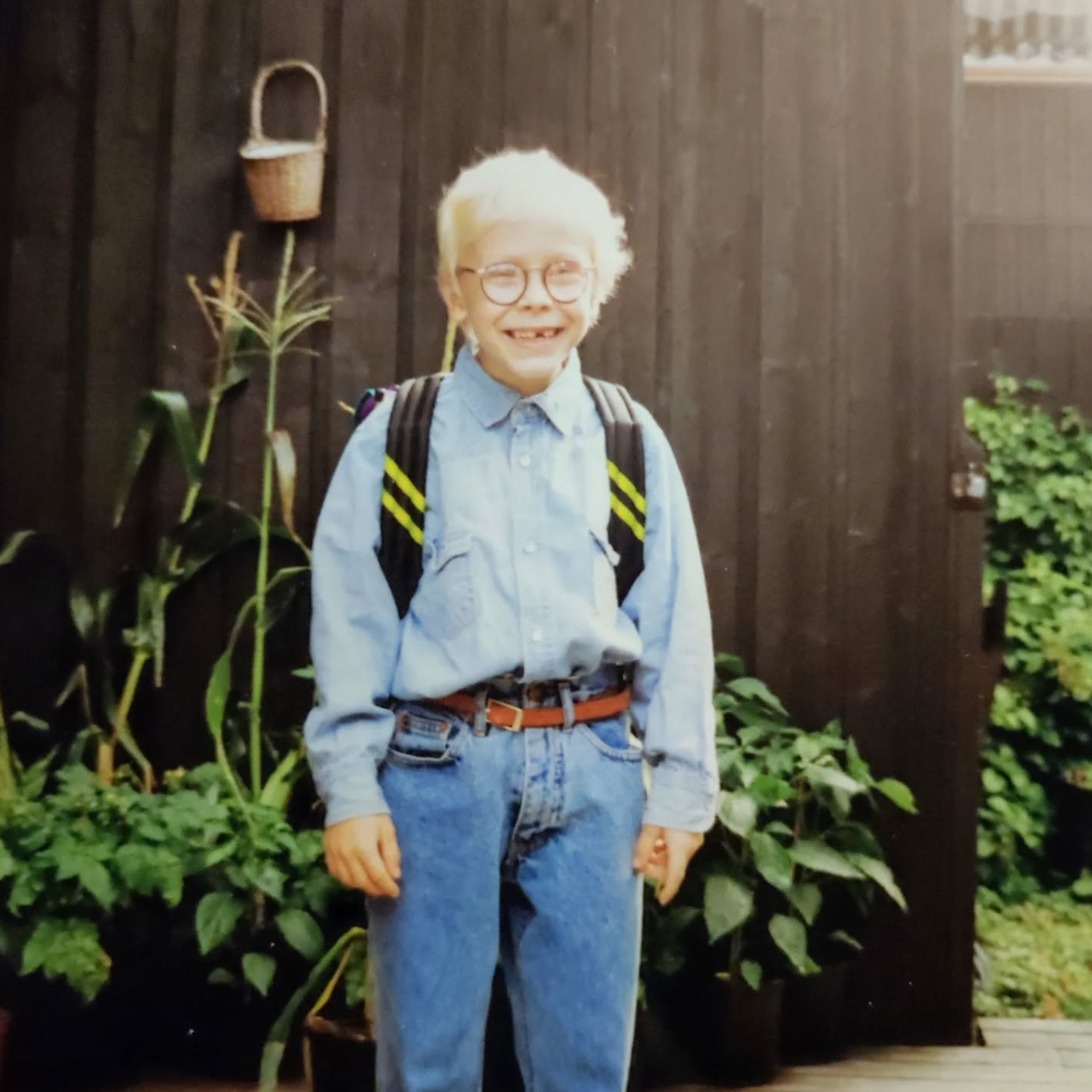 Ung jente med blond hår, iført blå skjorte og jeans, bærer ryggsekk og står foran en trevegg med planter rundt.