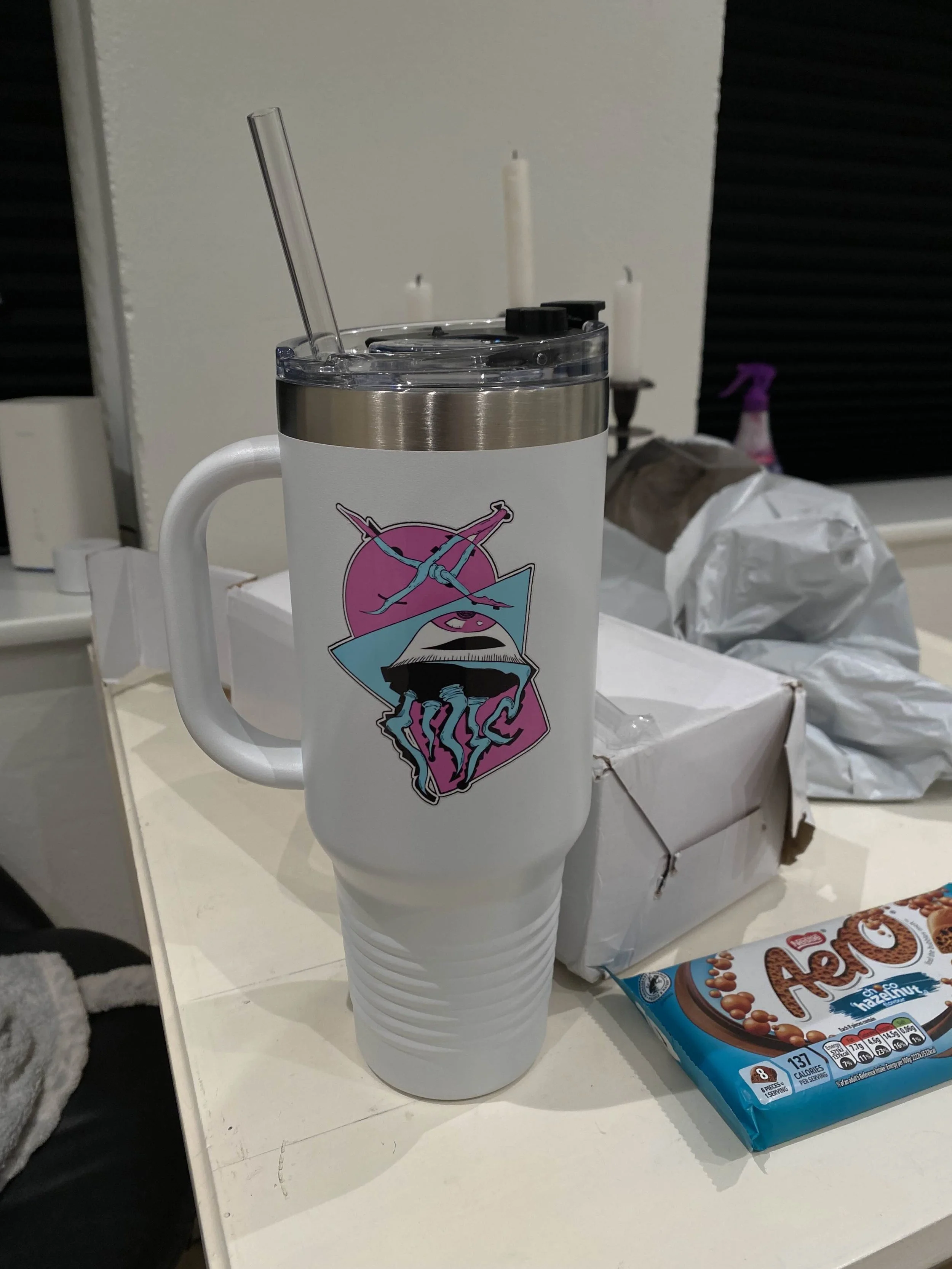 A white travel mug with a colorful graphic design featuring a pink circle, blue geometric shapes, and an illustration of a person with crossed arms. The mug has a clear lid with a drinking spout and a straw. The mug is placed on a cluttered white sur