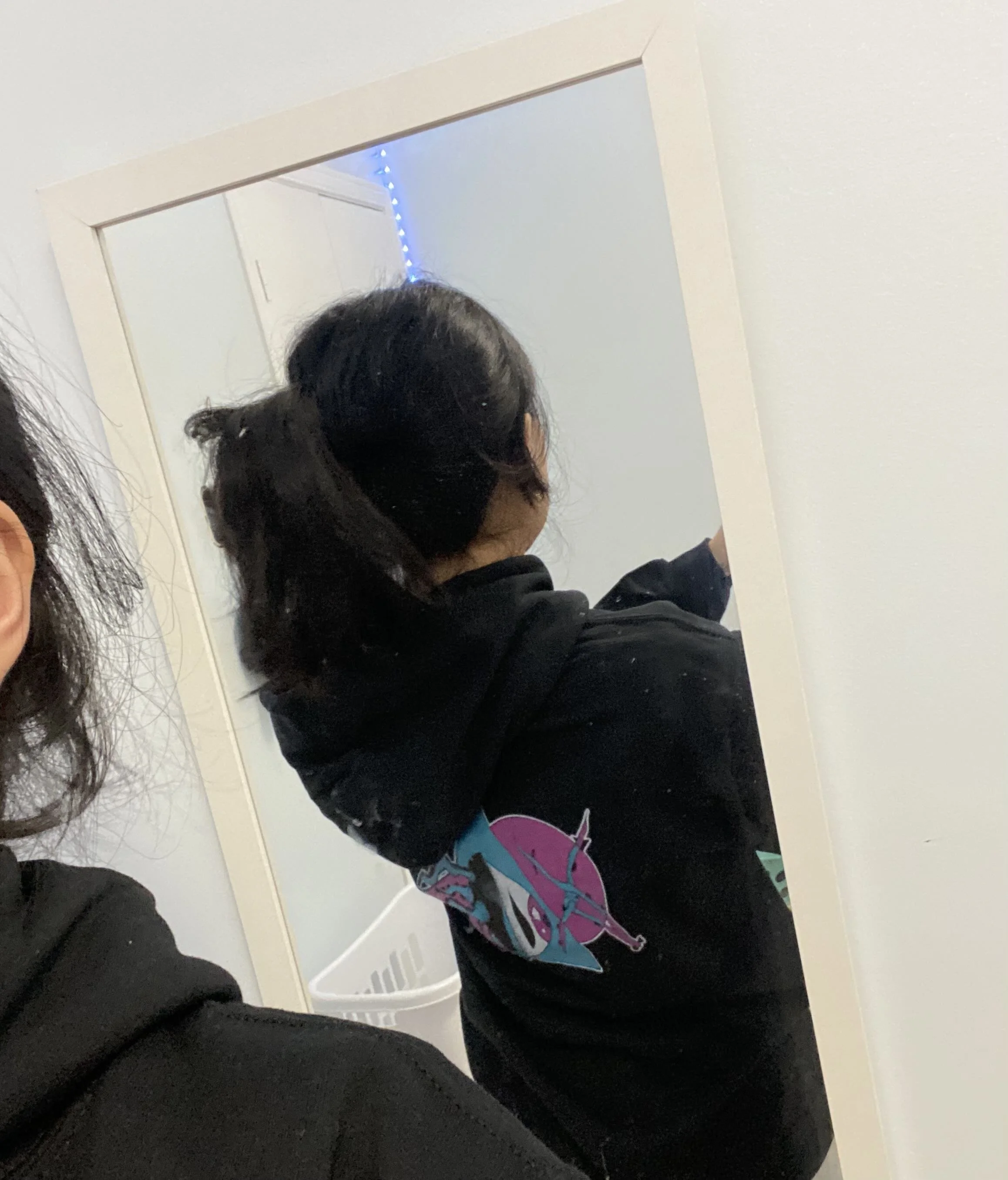 A person with black hair tied in a ponytail is standing in front of a mirror in a room with white walls. The individual is wearing a black hoodie with colorful graphic art on the front. The mirror reflects part of the room, including some lights and 