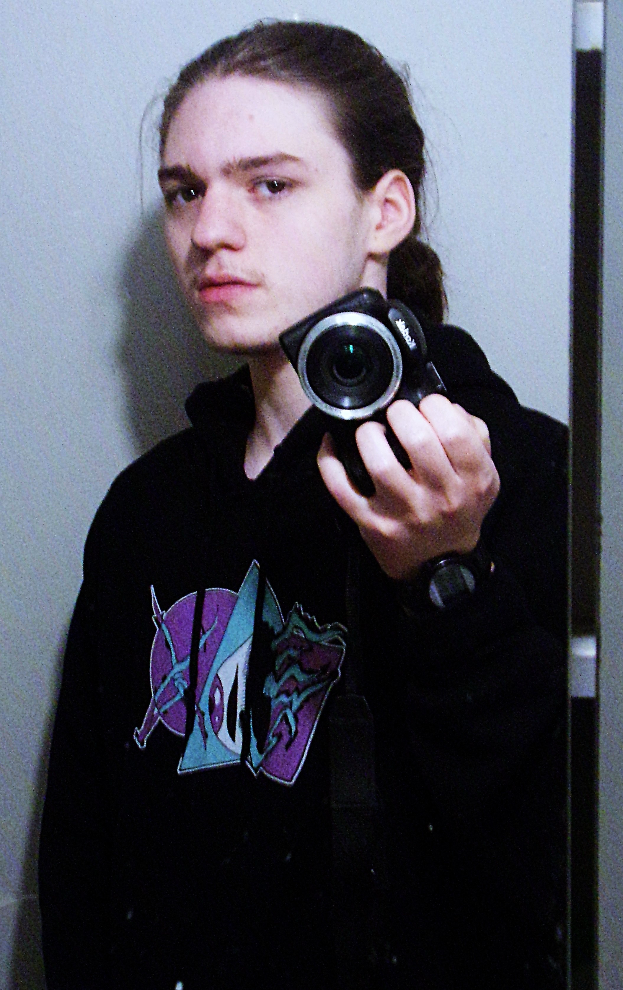 A young person with brown hair tied back, wearing a black hoodie with a colorful unicorn graphic, taking a mirror selfie with a digital camera.